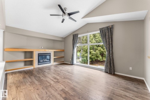 Vaulted ceiling with a ceiling fan, fireplace with a tile surround, built-in shelving, wood-finish flooring, and a large picture window - 2060 Brennan Crescent Nw, Edmonton, AB 