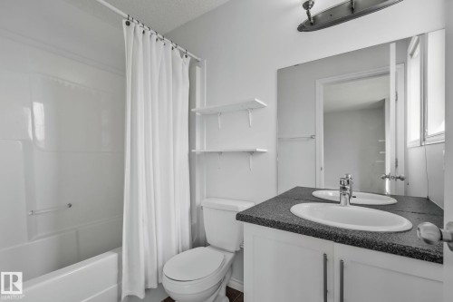 Bathroom featuring a white bathtub, toilet, and a vanity with a dark-finish countertop, two oval sinks, and a chrome faucet - 9523 Simpson Crest Nw, Edmonton, AB 