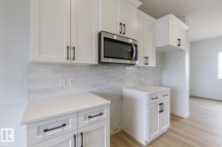 White cabinetry with matte black hardware, stainless steel microwave, light-toned countertop, subway tile backsplash, and light wood-finish flooring - 