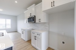 Contemporary kitchen featuring white cabinetry with black hardware, stainless steel microwave, light-toned wood-finish flooring, and a white subway tile backsplash - 