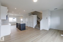 Open-concept living space featuring wood-finish flooring, a kitchen island with a basin sink and dark cabinetry, white upper and lower cabinetry, and a staircase with a carpeted runner and dark handrail - 