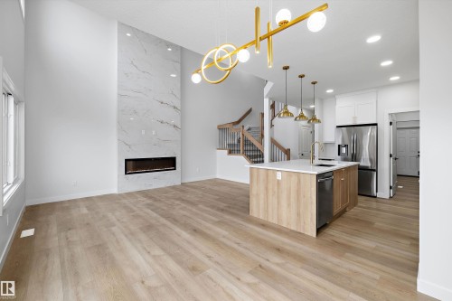 Spacious living area with wood-finish flooring, a two-story fireplace with a marble-style surround, and a modern gold chandelier - 1780 Crossbill Loop Nw, Edmonton, AB 