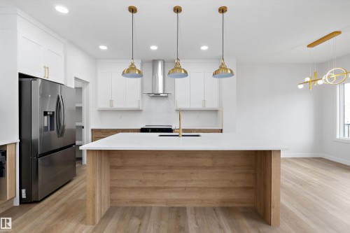 Kitchen featuring a central island with a white countertop and wood-finish paneling, stainless steel appliances, white cabinetry with gold-tone hardware, and wood-finish flooring - 1780 Crossbill Loop Nw, Edmonton, AB 