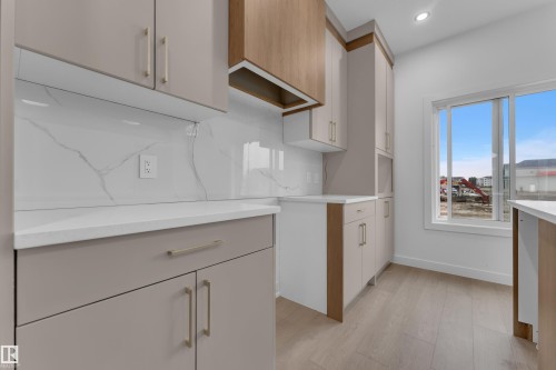 Cream-toned cabinetry with brass hardware, wood-finish accents, white stone countertops, a full-height backsplash, and wood-finish flooring - 265 Falcon Crescent, Leduc, AB 