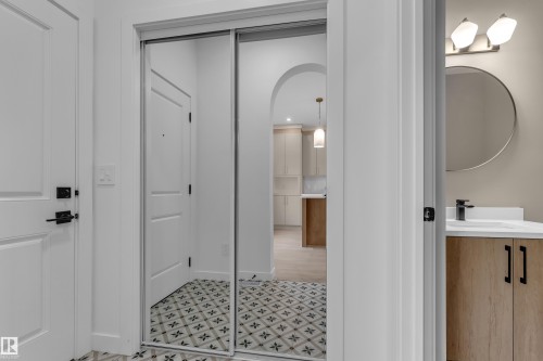 Custom tile flooring, mirrored closet doors, arched doorway, wood-finish vanity with black hardware, and a circular mirror with vanity lighting - 265 Falcon Crescent, Leduc, AB 