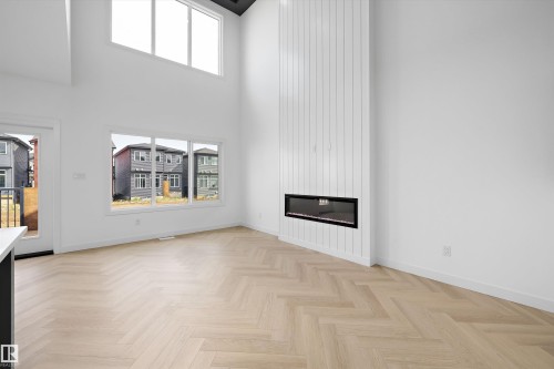 Double-height ceiling, herringbone wood-finish flooring, linear fireplace with vertical paneling, multiple large windows, and a glass-paneled exterior door - Edmonton, AB 