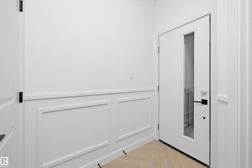 White paneled wainscoting, light wood-finish flooring in a herringbone pattern, contemporary white door with vertical glass insert, and matte black hardware - Edmonton, AB 