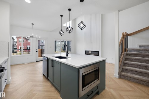 Open-concept living area featuring a kitchen island with integrated microwave, wood-finish flooring, a contemporary fireplace, and a carpeted staircase - Edmonton, AB 