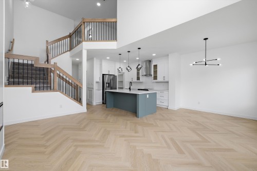 Open-concept living area with high ceilings, light wood-finish herringbone flooring, and a modern kitchen featuring an island with a light countertop and pendant lighting - Edmonton, AB 