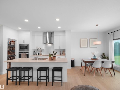 Open-concept kitchen and dining area featuring a light wood-finish flooring, white cabinetry, a large kitchen island with counter seating, stainless steel appliances, and a walk-in pantry - 22650 99A Avenue Nw, Edmonton, AB - Indoor