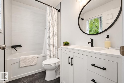 Integrated bathtub with white tile surround, white vanity with black hardware, black-framed oval mirror, matte black faucet, and durable dark-toned flooring - 1660 Tomlinson Common Nw, Edmonton, AB 