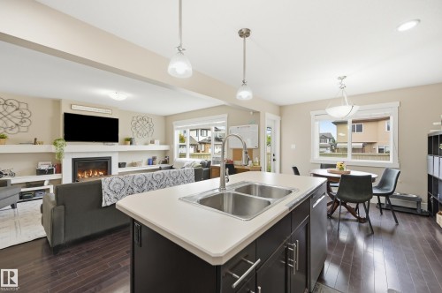 Open-concept living space featuring wood-finish flooring, a kitchen island with a double sink, a dining area with pendant lighting, and a living room with a fireplace framed by built-in shelving - 1564 36 Avenue Nw, Edmonton, AB 