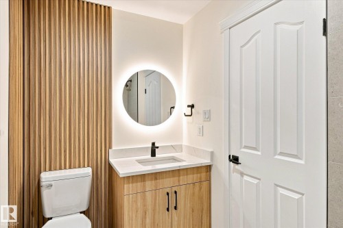 Bathroom featuring a wood slat accent wall, a floating vanity with a white countertop, a circular back-lit mirror, and a white paneled door with black hardware - 17325 85 Avenue Nw, Edmonton, AB 