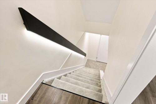 Modern staircase featuring a black angular handrail with integrated lighting, recessed wall lighting, carpeted treads, and wood-finish flooring - 17325 85 Avenue Nw, Edmonton, AB 