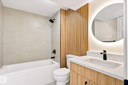 Bathroom featuring a white soaking tub, light-toned tile surround, wood-finish slat accent wall, illuminated round mirror, and a vanity with a white countertop and integrated sink - 17325 85 Avenue Nw, Edmonton, AB 