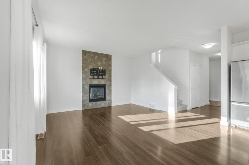 Wood-finish flooring throughout, featuring a fireplace with a stone-finish surround, staircase, and stainless steel refrigerator - 7832 Erasmus Wynd Nw, Edmonton, AB 