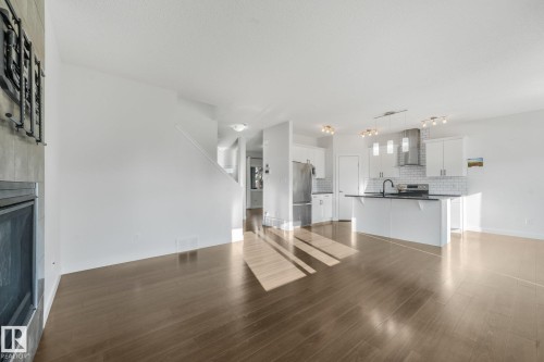 Spacious open-concept area featuring wood-finish flooring, a built-in fireplace, and a kitchen with white cabinetry, stainless steel appliances, and a central island - 7832 Erasmus Wynd Nw, Edmonton, AB 