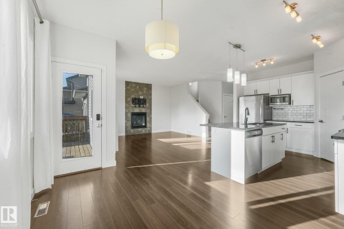 Hardwood-finish flooring, built-in fireplace with stone-finish surround, kitchen island with integrated sink, white cabinetry with stainless steel appliances, and a glass-paneled exterior door - 7832 Erasmus Wynd Nw, Edmonton, AB 