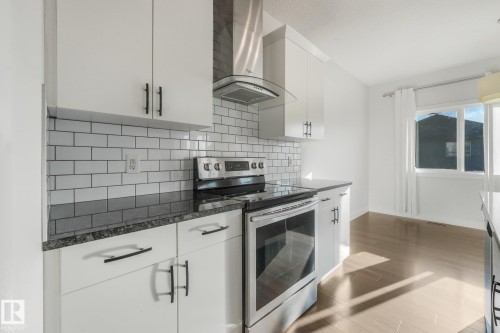 White cabinetry with matte black hardware, subway tile backsplash, stainless steel appliances, dark countertops, and wood-finish flooring - 7832 Erasmus Wynd Nw, Edmonton, AB 