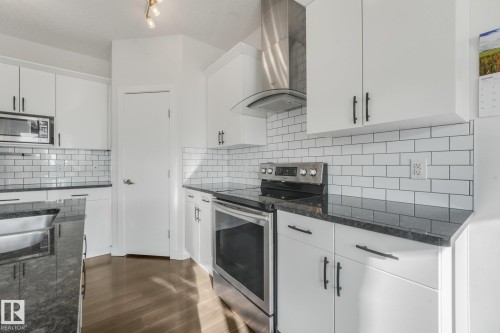 White cabinetry with matte black hardware, dark stone-finish countertops, stainless steel appliances, white subway tile backsplash, and wood-finish flooring - 7832 Erasmus Wynd Nw, Edmonton, AB 