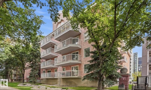 Multi-story building featuring stucco exterior, multiple private balconies with white railings, and landscaped grounds with mature trees - #107 10153 117 St Nw, Edmonton, AB 