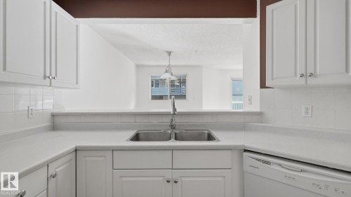 White cabinetry with silver hardware, dual basin stainless steel sink with a high-arc faucet, tiled backsplash, and white countertops - #107 10153 117 St Nw, Edmonton, AB 