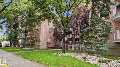 Building exterior with multiple balconies, mature trees, and a landscaped rock garden - #107 10153 117 St Nw, Edmonton, AB 