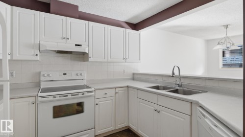 White cabinetry with silver-tone hardware, white tile backsplash, stainless steel double basin sink, and white appliances - #107 10153 117 St Nw, Edmonton, AB 