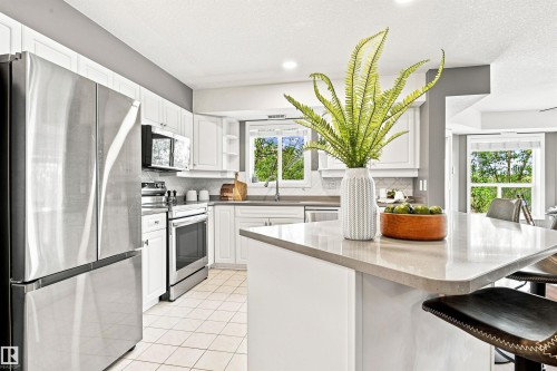 White cabinetry with sleek hardware, stainless steel appliances, light-toned countertop, tile flooring, and a kitchen island with seating - #432 6703 172 St Nw, Edmonton, AB 