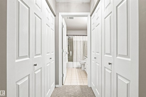 Hallway featuring bi-fold closet doors with dark hardware, leading to a bathroom with a ceramic tile floor, bathtub, and toilet - #432 6703 172 St Nw, Edmonton, AB 