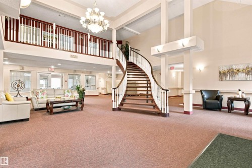 Elegant foyer featuring a grand curved staircase with wood-finish treads and metal railings, soaring ceilings with decorative coffers, a tiered chandelier, and expansive open floor plan with neutral carpet flooring throughout - #432 6703 172 St Nw, Edmonton, AB 
