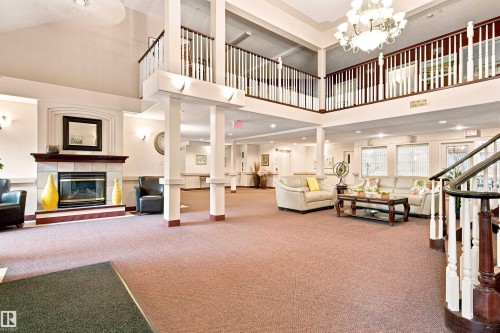 Spacious two-story lobby featuring a tiled fireplace, an elegant chandelier, and a grand staircase with white balusters and wood handrails - #432 6703 172 St Nw, Edmonton, AB 