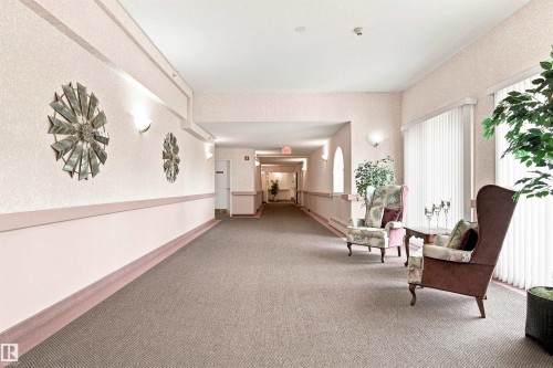 Carpeted hallway featuring wall sconces, decorative wall art, and vertical blinds on windows - #432 6703 172 St Nw, Edmonton, AB 