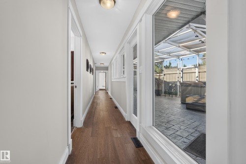 Wood-finish flooring throughout, featuring a long hallway with white trim, recessed lighting, and windows overlooking a covered outdoor patio with stamped concrete - 11603 72 Avenue Nw, Edmonton, AB 