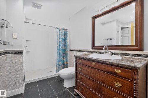 Dark tiled flooring, wood-finish vanity with a mounted sink, large framed mirror, and a white shower stall with built-in shelving - 11603 72 Avenue Nw, Edmonton, AB 