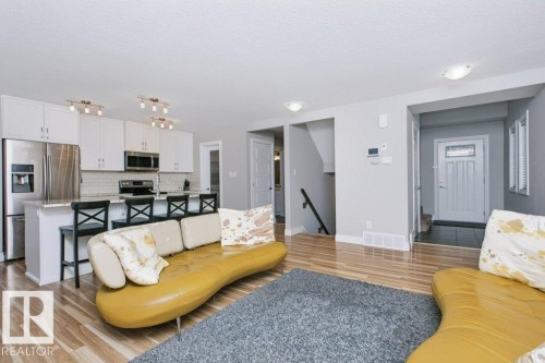 Open-concept living area featuring wood-finish flooring, a contemporary kitchen with white cabinetry, stainless steel appliances, a kitchen island with seating, and track lighting fixtures - 3496 Weidle Way Sw, Edmonton, AB - Indoor