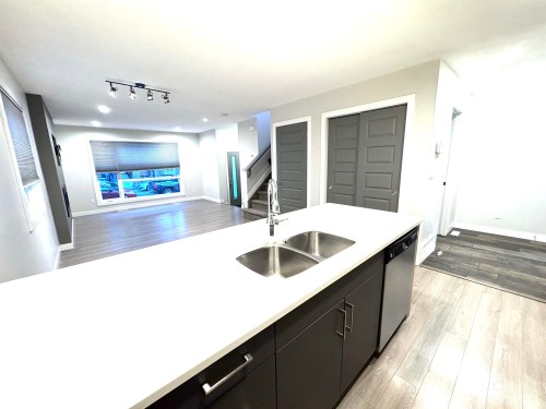 Open-concept living space featuring wood-finish flooring, a large window, a track lighting fixture, and a kitchen island with a double stainless steel sink - 12046 89 Street Nw, Edmonton, AB 