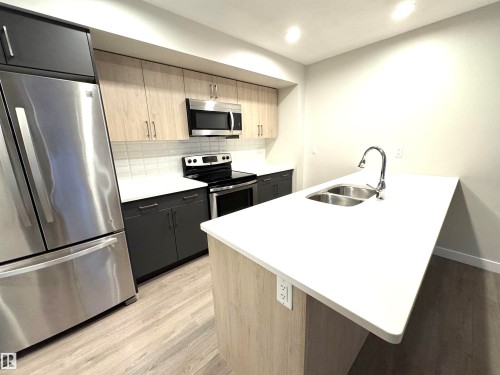 Modern kitchen featuring stainless steel appliances, dual-tone cabinetry, wood-finish flooring, a white countertop island with an integrated double sink, and a tiled backsplash - 12046 89 Street Nw, Edmonton, AB 
