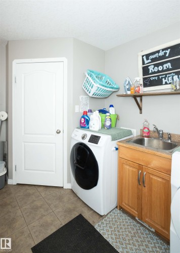 Utility room featuring a front-load washing machine, tile flooring, and a single-basin utility sink with a wood-finish cabinet - 6003 50 Ave, Barrhead, AB 