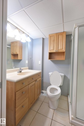 Light wood-tone vanity with integrated sink, coordinating wall-mounted cabinetry, tile flooring, and a glass-enclosed shower - 8127 7 Avenue Sw, Edmonton, AB 