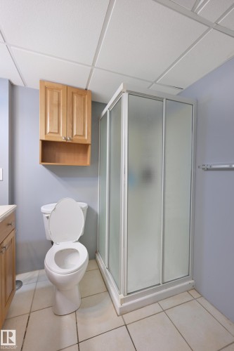 Walk-in shower with frosted glass enclosure, white ceramic tile flooring, light wood-finish cabinetry, and a white toilet - 8127 7 Avenue Sw, Edmonton, AB 