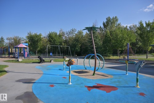 Splash pad with aquatic-themed features, adjacent to a playground with swings and slides, surrounded by mature trees - 8127 7 Avenue Sw, Edmonton, AB 