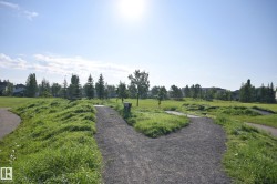 Gravel pathways traversing a large, open green space with varied topography, bordered by mature trees and residential structures in the distance - 