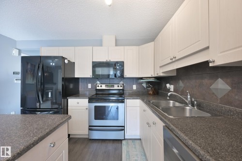 Kitchen featuring white cabinetry, dark speckled countertops, a dark tile backsplash, a double basin stainless steel sink, and wood-finish flooring - 8127 7 Avenue Sw, Edmonton, AB 