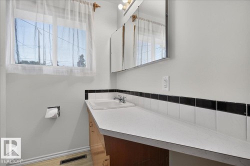 Vanity with integrated sink, expansive countertop, white wall tile with a black accent border, dual mirrored medicine cabinets, and a window providing natural illumination - 6104 92 Avenue Nw, Edmonton, AB 