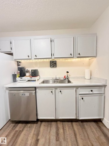 White cabinetry with dark hardware, stainless steel double basin sink, light-toned countertop, stainless steel dishwasher, and wood-finish flooring - #89 2703 79 St Nw, Edmonton, AB 