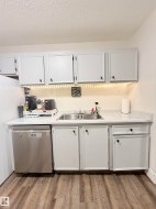 White cabinetry with dark hardware, stainless steel double basin sink, light-toned countertop, stainless steel dishwasher, and wood-finish flooring - 