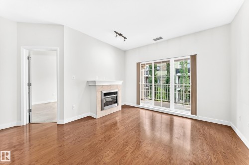 Spacious room featuring wood-finish flooring, a contemporary fireplace with a tile surround and mantel, and a large sliding glass door with a balcony - #307 1003 Gault Bv Nw, Edmonton, AB - Indoor Photo Showing Other Room With Fireplace