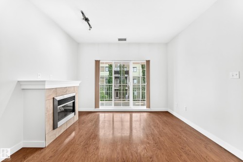 Spacious interior featuring wood-finish flooring, a contemporary fireplace with a tile surround and white mantel, and a large sliding glass door - #307 1003 Gault Bv Nw, Edmonton, AB - Indoor With Fireplace