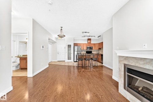 Spacious living area featuring wood-finish flooring, a tiled fireplace with a white mantel, and an open-concept kitchen with wood cabinetry and stainless steel appliances - #307 1003 Gault Bv Nw, Edmonton, AB - Indoor Photo Showing Living Room With Fireplace